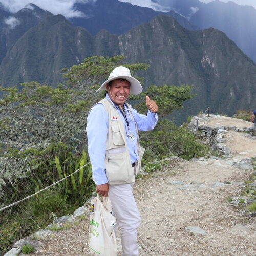 A happy and welcoming Peruvian tour guide in bright clothes wearing a beige hat and giving the thumbs up