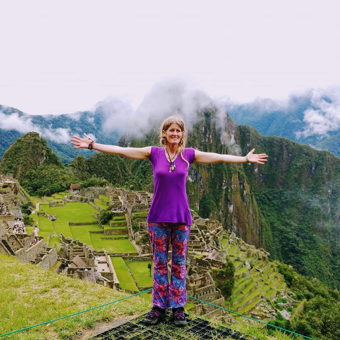 A woman dressed in colorful clothes standing on a mountainside in front of a mountain and green grass with open arms and a welcoming smile